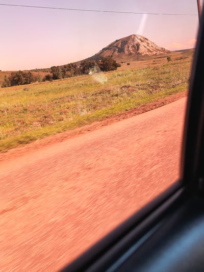 The image is a landscape photograph taken from inside a vehicle, likely looking out of a car window. The foreground is dominated by a reddish-orange, blurred road surface, indicating motion. Beyond the road, a grassy field stretches out, dotted with sparse vegetation and small trees. In the mid-ground, a prominent, cone-shaped hill or mountain rises, its summit appearing lighter in color, possibly due to exposed rock or a different soil composition. Darker green trees and foliage cover the lower slopes of this prominent landform. The sky above is a soft, pale pink or purple hue, suggesting it might be sunrise or sunset. A thin, dark line, possibly a power line or telephone wire, is visible stretching horizontally across the upper portion of the sky. The overall scene appears to be rural and natural, with no visible human activity or structures, other than the implied vehicle from which the photo is taken. The provided location of Karaiganovo, Russia, places this natural landscape within that specific geographic context.