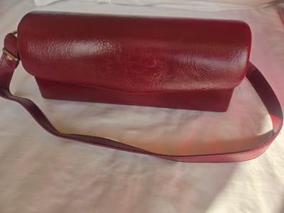 A red leather handbag with a single strap lies on a white textured surface. The handbag is rectangular with a curved flap closure and a textured leather finish. A thin, adjustable strap extends from the side of the bag, with a gold-colored buckle visible. The background is a white, rumpled fabric that casts soft shadows, suggesting indoor lighting. There are no people or other objects visible in the image.