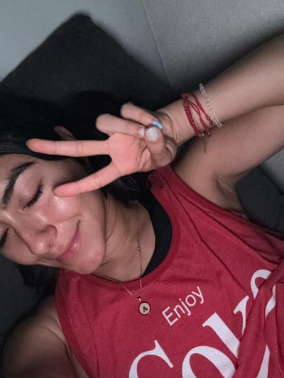 In the image, a person is lying down, with their face tilted upwards and their eyes closed. They are wearing a red tank top with the word "Enjoy" visible on it, along with a partial Coca-Cola logo. A gold necklace with a circular pendant is also visible. The person's right hand is raised, with two fingers extended in a peace sign gesture. They are also wearing several bracelets on their wrist, including a red beaded one and a clear beaded one. The background appears to be a dark, textured surface, possibly a couch or pillow, against a gray wall. There is no indication of the city of Imatra, Finland in the image.