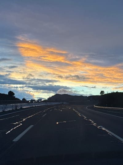 The image depicts a multi-lane highway scene at dusk or dawn, specifically located near Lorca, Spain. The sky dominates the upper portion, featuring distinct bands of orange and yellow-lit stratocumulus clouds against a backdrop of grey and light blue. In the midground, a dark, silhouetted mountain range is visible against the horizon. The highway, dark in tone, occupies the lower and central portions of the frame, showing two apparent lanes in the foreground with white dashed and solid line markings. Patches of reflective moisture are visible on the road surface. To the left, a concrete barrier system separates the road from an elevated embankment with sparse vegetation. To the right, a metallic guardrail curves alongside another vegetated embankment, with a solitary tree silhouetted against the sky further up the hill. Several vehicles with illuminated headlights are discernible in the distance, traveling on the highway. No individuals are visible.