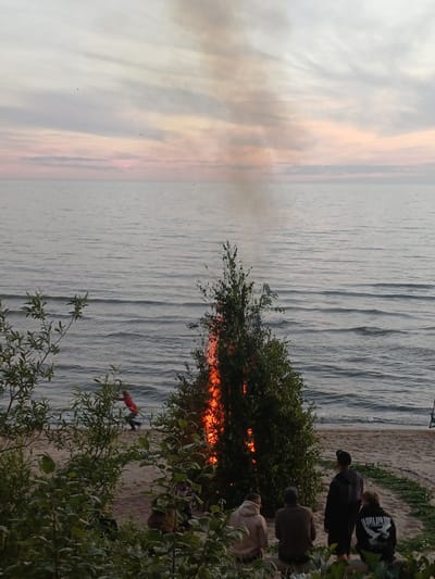 The image captures a serene beach scene at dusk in Vārzas, Latvia. The sky is awash with soft, pastel hues of orange and pink, indicative of a sunset. The calm, rippling water of a lake or sea stretches across the frame, meeting a sandy beach dotted with patches of green vegetation.

Several people are present, observing an event taking place near the water's edge. They are casually dressed, suggesting a relaxed atmosphere. Some are standing, others are crouching or sitting, engrossed in what is happening.

The focal point of the activity is a bonfire, or some other fiery spectacle, that illuminates the foliage of a nearby tree with a bright orange glow. Smoke rises from the fire, partially obscuring the scene and adding a touch of drama. A person in a bright red outfit is visible near the fire, possibly interacting with it or observing from a closer vantage point.

Among the people, one individual is wearing a black hoodie with white text. Though slightly obscured, the text appears to say "WORLDWIDE". The overall mood of the image is one of quiet contemplation and communal gathering, with the natural beauty of the sunset and the captivating fire creating a memorable experience.