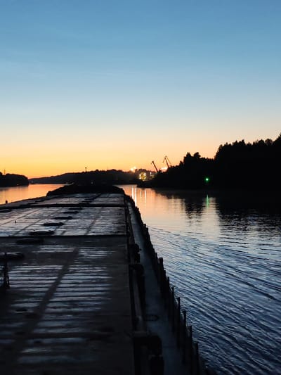 The image depicts a wide river at dusk, with a clear gradient of colors in the sky, transitioning from a pale orange on the horizon to a deep blue overhead. On the left side of the frame, a large, flat barge or pier extends into the water, its surface appearing wet and reflecting the fading light. The barge is dark and industrial, with visible patterns of raised elements and circular indentations. Along the right edge of the barge, a railing or low fence runs the length of its visible portion.
The water is calm but shows ripples and waves, suggesting movement, possibly from a vessel or current. The right bank of the river is densely forested, appearing as a dark silhouette against the twilight sky. In the distance, along the far bank, industrial cranes are visible, illuminated by artificial lights, suggesting the presence of a port or industrial area. A single green light is visible on the water, likely a navigational marker. The overall atmosphere is serene and quiet, characteristic of the end of the day. The location is Annenskii Most, Russia.
