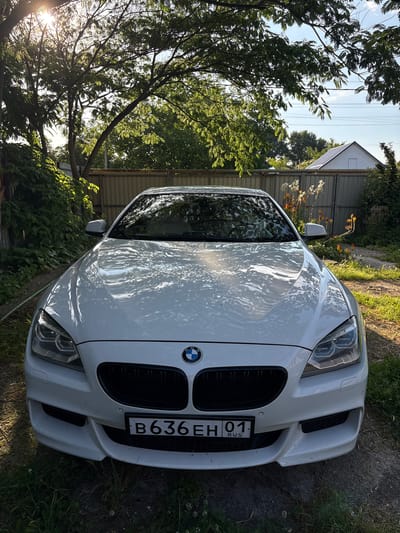 The image shows the front of a white BMW sedan parked outdoors in a grassy area. The car is the main subject, with its grille, headlights, and license plate clearly visible. The license plate reads "B636EH 01 RUS". The scene is set in what appears to be a rural or suburban backyard, with trees, bushes, and a fence in the background. A building with a white roof and metal siding is also visible behind the trees. The sun is shining, casting dappled light on the car and the surrounding foliage, suggesting it is daytime. The lighting and shadows indicate a sunny day, possibly late afternoon or early morning due to the low angle of the sun. There are no people visible in the image. The overall impression is a still shot of a car in a natural, outdoor setting.
