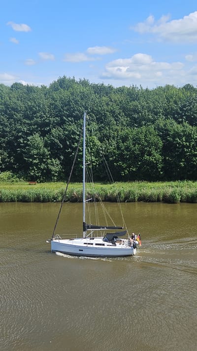 This is a vertical, eye-level, medium shot photograph taken on a sunny day in Kudensee, Germany. The image captures a white sailboat with a tall mast, moving left to right on a wide, calm canal. The boat has a dark-colored sail cover, and two people are visible on the deck. One person is steering, and another is standing near the stern. A German flag is attached to the stern of the boat.

The canal's water is a murky brown-green color, with visible ripples on the surface created by the boat's movement. The canal's edge is lined with tall, green grass and reeds. Beyond the grassy bank, a dense forest of lush green trees forms a thick wall, with a narrow dirt path visible running along the treeline. A single wooden bench is positioned on the path, facing the canal. The sky is a bright blue with scattered white, fluffy clouds.