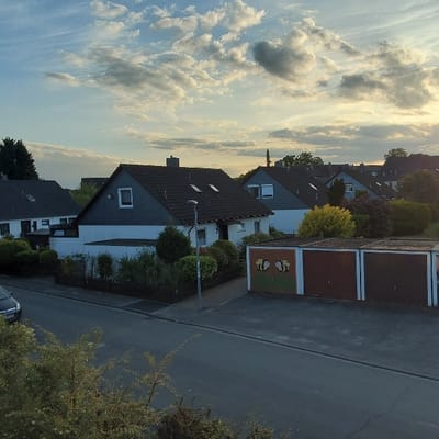 The image shows a residential street in Garbsen, Germany, with a row of houses and a garage. The houses are white with dark gray roofs. The garage has brown doors and a white wall with a mural depicting a beehive. The street is asphalt and bordered by a green lawn. The sky is blue with white clouds. There is a car parked in the street. There are trees and shrubs on the lawn. There are no people present. The coordinates of the location are 52.415 latitude and 9.591 longitude.