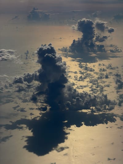 The image presents an aerial view, likely taken from a high altitude such as an airplane or satellite, of a cloudscape with a body of water or land reflecting light visible in between the clouds. The clouds themselves vary in density, with some appearing as thick, dark masses and others as thin, dispersed patches. The overall environment suggests either a coastal area or an open expanse of water, based on the reflections and cloud patterns. Without further context, pinpointing the precise location in Taoyuan City, Taiwan, is impossible.