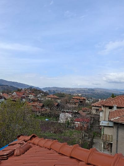 An elevated, panoramic view of the settlement of Krupnik, Bulgaria, is presented under a light blue sky with scattered cumulus clouds. The immediate foreground features a terracotta-tiled rooftop, with several tiles stacked and a section covered by a blue synthetic sheet. The midground reveals a cluster of residential structures, predominantly with red or orange ceramic tile roofs, interspersed among numerous trees and cultivated plots. The vegetation includes deciduous trees exhibiting early spring foliage, some with light green leaves and others displaying clusters of pink-purple blossoms. The background shows undulating terrain comprising forested and less vegetated hills and mountains receding into the distance. No human figures are visible within the depicted scene.