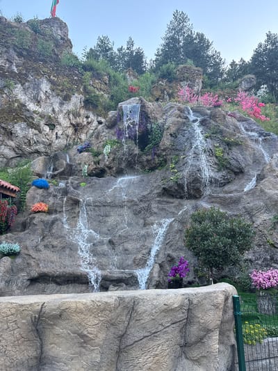 The image depicts a vertical rock formation with a waterfall cascading down its face, adorned with a vibrant display of artificial flowers in various colors including pink, purple, blue, and orange. The rock face is textured, with crevices and ledges, and patches of moss and greenery cling to its surface. Several trees with dark green foliage are visible at the top of the rock. In the background, there is a hint of a fence and a grassy area, suggesting an outdoor setting. A flag with red, white, and green bands is partially visible on the left side of the rock. The sky appears overcast, indicating a potentially cloudy or misty day. The overall impression is one of a decorative, landscaped feature.