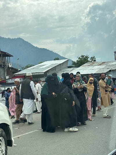 In Cherwan, India, a diverse group of people stands on a street under a cloudy sky. The foreground features a white car, partially visible on the left, and a paved road with white lines. Several individuals are walking or standing. Men are dressed in traditional attire, including kurtas and waistcoats, while women wear hijabs and burqas in various colors. One man carries a baby. Behind the people, buildings with corrugated metal roofs are visible, suggesting a commercial or residential area. In the background, a large, tree-covered mountain looms under the overcast sky.