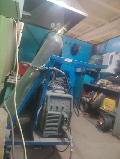 The image depicts a cluttered workshop environment, possibly a garage or industrial space. In the foreground, a welding machine is visible, connected to cables. To the left, a large, green gas cylinder with gauges is positioned on a blue metal frame. The cylinder appears to be secured by a chain. In the background, there are various pieces of equipment, shelves filled with items, and what appears to be a blue metal enclosure or machine. The floor is concrete and scattered with debris. The overall impression is one of an active or recently used workspace. The location is stated as Пліскі сельскі Савет, Belarus. There are no people visible in the image.