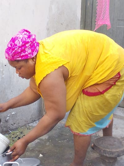 A woman in a bright yellow dress and pink headwrap leans forward. She appears to be cooking outside. Her left hand holds a metal pot over a larger covered pot. Behind her is a concrete wall, a small wooden stool, and a doorway with a pink net hanging near it. Water is on the ground around her feet, and the overall scene appears to be in a residential or outdoor kitchen setting.