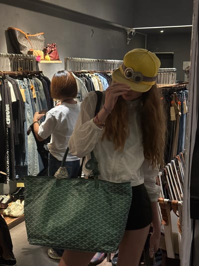 The image captures a retail environment, likely a vintage clothing store, with racks of garments visible. In the foreground, a person wearing a yellow cap with novelty goggles and a white long-sleeved shirt is holding a large, dark green tote bag with a patterned design. Another person with short brown hair is visible in the mid-ground, appearing to browse clothing on a rack. The store interior features a dark gray wall and shelves displaying various bags. The overall impression is that of a casual shopping scene. This scene is located in the Xuhui District, China.