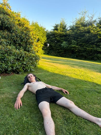 A shirtless individual with light brown hair is lying on their back on a grassy lawn in front of a large bush and some evergreen trees. They are wearing black shorts and have a black backpack underneath their head. The scene appears to be a residential backyard, possibly in the city of Mordarka, Poland. The sun is shining, casting long shadows across the lawn, indicating it is late afternoon or early evening. The individual's arms are outstretched to their sides, and their legs are slightly bent. The overall impression is one of relaxation or repose.