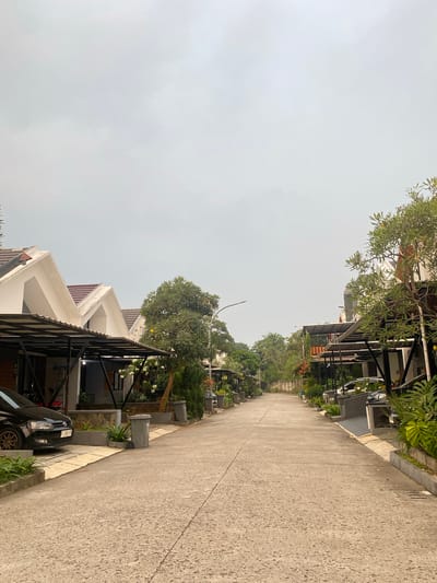 The image depicts a straight, paved road in a residential area in Cibinong, Indonesia. The sky is overcast and a light grey. On both sides of the road are houses with modern, angular roof designs, characterized by sharp peaks and white facades. Each house features a covered carport with black metal supports.
To the left, a black Volkswagen sedan is parked under one of the carports. A grey trash bin is situated on the pavement near the car. Several potted plants are visible along the property lines.
To the right, another black car is parked under a carport. Lush green foliage, including trees and large-leafed plants, lines the properties.
The road itself is made of concrete and appears to be in good condition. The overall impression is of a well-maintained, quiet suburban neighborhood. There are no people visible in the image.