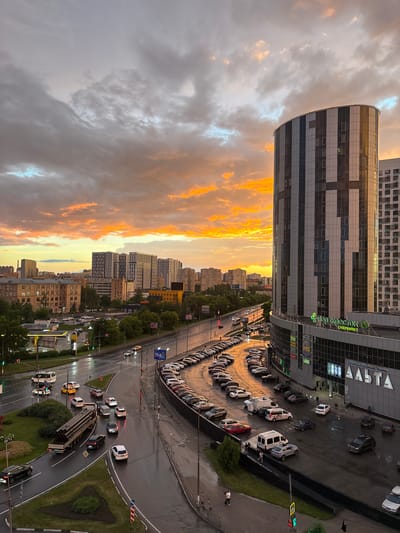 This is an aerial, high-angle, vertical, color photograph of a cityscape at dusk. The sky is dramatic, with a mix of dark gray clouds and vibrant orange and yellow sunset hues. In the foreground and midground, a multi-lane road with wet asphalt curves through a semi-urban environment. Several cars, including a white van, a yellow sedan, and a white SUV, are visible on the road. A large truck carrying pipes is moving away from the camera. There is a car dealership with a yellow awning and a gas station on the left side of the road. Further into the midground, a modern, curved, glass-and-metal high-rise building dominates the right side of the frame. The building has a distinctive pattern of dark and light vertical panels and displays a sign that reads "ПЕРЕКРЕСТОК" in green lettering above "СУПЕРМАРКЕТ". Adjacent to this building is another tall, white residential building. In the background, a dense cluster of multi-story residential buildings and some older, lower-rise apartment blocks are visible, receding into the distance under the colorful sky. The environment appears to be recently rained on, given the wet roads and reflections. There are a few scattered pedestrians visible in the image, mostly near the commercial building. The scene depicts a bustling urban area during a dramatic sunset.