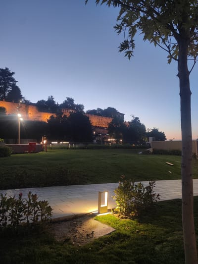 This media file captures an outdoor scene at dusk, likely in Osmangazi, Turkey, given the context. The main subjects are a well-maintained park with manicured grass, a paved walkway, and low-lying bushes. In the background, a large, ancient stone wall, illuminated by warm orange lights, stretches across a hillside. This wall appears to be part of a historical fortification or structure, possibly a castle or citadel, overlooking the park. Dense foliage, including trees, grows along the top of the wall and on the hillside, silhouetted against the fading twilight sky.

Several modern elements contrast with the historical backdrop. Sleek, rectangular garden lights are strategically placed along the walkway, casting a soft yellow glow onto the pavement and surrounding grass. A tall, narrow lamppost with multiple lights is visible closer to the stone wall, providing further illumination. The presence of what looks like a red mailbox near the lamppost suggests a public or residential area. A young tree trunk is prominently featured in the foreground on the right side of the frame.

The time of day is clearly indicated by the twilight sky, which transitions from a deep blue at the top to a lighter hue closer to the horizon, where a faint glow suggests the last vestiges of sunset. The weather appears calm and clear. The overall mood is tranquil and serene, with the combination of natural elements, historical architecture, and modern lighting creating an inviting atmosphere.

Visible text includes "LOUNGE & CAFE" illuminated on a sign situated behind a fence and greenery, suggesting a commercial establishment is located within or near the illuminated wall structure. The scene evokes a sense of peace and the blending of history with contemporary life.