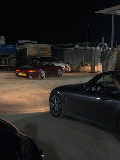 The image captures an outdoor scene at night, likely in a parking or industrial area, as suggested by the presence of multiple vehicles, trucks, and shipping containers. The perspective is from slightly above, looking down towards the ground.

In the immediate foreground, a dark gray car, possibly a convertible or coupé, is partially visible on the right side. Its driver's door is open, and a person's head and shoulders can be seen in the driver's seat, suggesting they are either entering or exiting the vehicle. The car features multi-spoke alloy wheels.

Further into the scene, towards the left, a silver hatchback is parked, followed by a dark red sedan. The license plate on the dark red sedan is clearly visible, displaying "58-37-29" and the word "ישראל" (Israel), confirming the location context as Israel. In the bottom right corner, a portion of another dark-colored car is visible.

The background on the left features various industrial elements, including what appear to be trailers or truck beds with reflective safety markings, and several blue shipping containers. One container has faded white text including "153-4815369". Overhead, near some basic structures, electrical wires and conduits are noticeable. The setting is illuminated by artificial light sources, casting shadows on the ground and vehicles. The sky is completely dark, indicating it is nighttime.