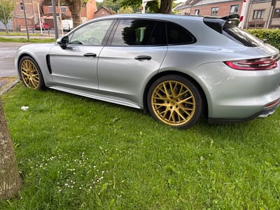 A silver Porsche Panamera Sport Turismo is parked on a patch of green grass with small white flowers. The car features gold-colored wheels and black door handles. The background shows a street scene in Melreux, Belgium, with brick buildings, a white van, and red car visible across the street. A tree trunk is positioned on the left edge of the frame.