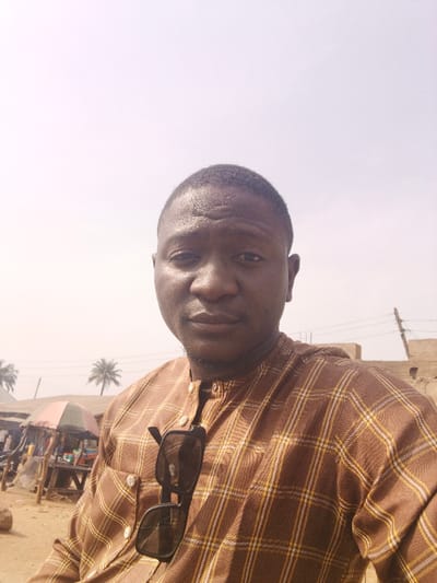 A medium shot of a man wearing a brown and yellow plaid shirt with sunglasses hanging on the front. The background shows an outdoor market setting with stalls, umbrellas, and palm trees under a hazy sky. He is looking directly at the camera. The composition is a selfie-style photo.