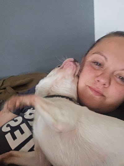 A selfie shows a woman holding a light-colored dog. The woman has fair skin, brown eyes, and dark hair pulled back. She is wearing a dark t-shirt with white lettering. The dog is pressed against her face and chest. The background is a plain wall, with a gray lower portion and a white upper portion. The location in the city of Hem, France, cannot be determined from the image.