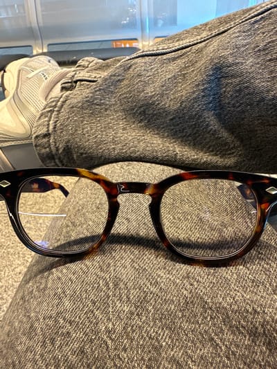 This close-up image, captured in Weeze, Germany, features a person's leg clad in dark grey denim jeans and a white athletic shoe, with a pair of tortoiseshell eyeglasses resting prominently on the fabric. The denim displays a distinct twill weave texture with subtle folds. On the lower left, a white sneaker with grey stripes and accents is visible, though its brand logo is indistinct due to blurriness.
The eyeglasses, the central subject, boast stylish dark brown and amber mottled frames. A small, silver-colored diamond-shaped inlay decorates the upper corner of the right lens frame. On the bridge connecting the two lenses, a unique, stylized symbol resembling a wave or an 'M' is etched. The clear lenses show a slight blue reflection, possibly indicating blue light filtering or an anti-reflective coating.
The background is softly blurred, revealing light-colored, linear structures, possibly shelving or a part of a cabinet, suggesting an indoor setting. The image captures a moment of stillness, with the items appearing to be temporarily placed. No specific activity is taking place, nor are any emotions discernible. The lighting appears to be ambient indoor light, but the time of day is not evident. No explicit text is clearly legible, apart from the stylized symbol on the eyeglasses.