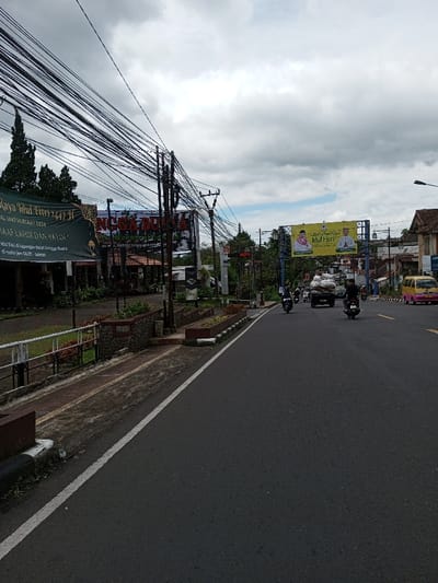 The image depicts a street scene in the city of Taohom, Laos, under a predominantly overcast sky with scattered grey clouds. An asphalt road, visible in the foreground and extending into the distance, features a solid white line delineating its left edge.
On the left side of the road, a raised concrete median or sidewalk runs parallel, equipped with low stone walls, planter boxes, and a white metal guardrail. Directly above this area, a dense network of black utility cables crisscrosses horizontally, supported by multiple vertical utility poles.
Further left, beyond the median, several structures and signs are present. A large green banner displays partially visible white text, including "RAYA IDUL FITRI 1447 H" and "MAAF LAHIR DAN BATIN." Adjacent to this, a prominent sign for "NAGA DUNIA" and "JENDELA ALAM" is visible, along with text referring to "www.hotelsangria." A red "HOTEL" sign is also present further down the street on the left. Tall, slender conifer trees and other greenery are visible in the background on this side.
Vehicular traffic is present on the road. In the mid-ground, three motorcycles are in motion: two in the left lane, and one in the right lane. Each motorcycle has at least one individual rider. A dark-colored flatbed truck or lorry, heavily loaded with numerous white sacks or bundles, travels in the right lane. To the far right, a yellow and pink minivan is visible.
In the background, a large yellow billboard spans horizontally across the road, displaying images of two men and the text "Selamat Hari Raya Idul Fitri." Buildings with varying roof types line the street in the distance.
