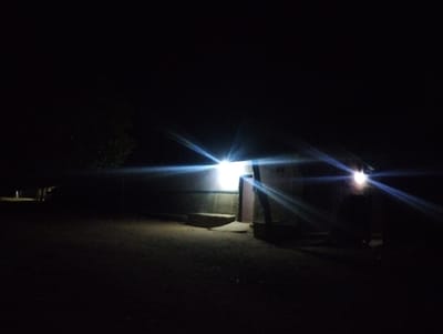 The image depicts a dimly lit nighttime scene in Bajoga, Nigeria, at the coordinates 10.852° N, 11.426° E. There is a small, one-story building illuminated by a single bright light. The light source is a bare bulb, casting a strong, focused beam. In the foreground, two figures are standing in front of the structure. Their features are obscured by the shadows. The surroundings are obscured by darkness, but there are indications of a dirt ground and some vegetation. The air is still and quiet. The scene suggests a typical rural setting in Nigeria.