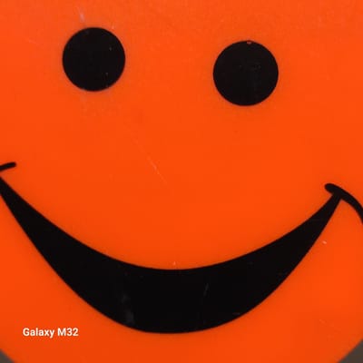 The image is a close-up shot of a smiley face drawn on an orange surface. The smiley face consists of two black circles for eyes and a curved black line for a mouth. In the bottom left corner of the image, the text "Galaxy M32" is visible. The surface appears to be plastic and shows some minor scratches and imperfections. The lighting is even, with no harsh shadows. The image does not contain any information about a specific location in Olsztyn, Poland.