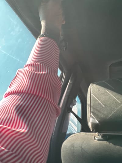 The image depicts the interior of a vehicle, presumed to be in Dokwa, Nigeria. A dark-skinned arm, dressed in a red and white vertically striped long-sleeved shirt, extends upwards towards the vehicle's light gray headliner. A dark beaded wristband is visible on the wrist, from which a small, dark, decorative object (possibly a skull figurine) hangs. Through the vehicle's window, a clear blue daytime sky is visible to the left. On the right, a dark gray car seat with faint diagonal striped patterns is partially visible, illuminated by direct sunlight. A black seatbelt and its attachment mechanism are also present on the dark gray pillar to the right of the arm.