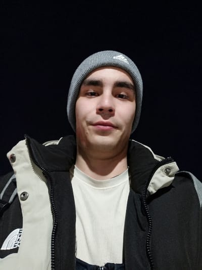 The image displays a head-and-shoulders portrait of a male individual, identified as being in the city of Yukhta, Russia. The subject is a young adult with a light-to-medium complexion, dark hair, and brown eyes, looking directly at the camera with a neutral expression. He is illuminated by an overhead light source against a uniform dark background, indicating a nighttime or low-light environment.

The individual wears a grey knitted beanie with a white Adidas logo prominently displayed on the front. He is dressed in a light beige or off-white ribbed long-sleeved shirt underneath an open, multi-paneled winter jacket. The jacket is primarily black, with light beige or off-white sections across the upper chest and shoulders, and grey segments on the sides and sleeves. A white stylized logo, resembling "THE NORTH FACE" with a mountain graphic, is visible on the left chest area of the jacket. The jacket features a black central zipper and silver-colored snap buttons on the collar flaps. A faint reddish mark or discoloration is observed on the right side of the subject's jawline/neck. No other individuals or objects are discernible in the background.