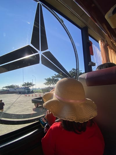 The image depicts the interior of a vehicle, likely a bus, looking outward through a large window featuring the prominent Mercedes-Benz three-pointed star logo integrated into its design. The exterior scene, set in Sumber Suko, Indonesia, shows a sunny day over an asphalt lot or parking area. Several large box trucks and flatbed trucks are parked, alongside a dark-colored pickup truck with an open bed. Orange traffic cones are arranged on the asphalt, and tall light poles are visible. Green trees line the perimeter of the lot, with a hazy horizon suggesting distant fields or foliage under a clear blue sky.

Inside the vehicle, a person is seen from the back in the foreground, wearing a wide-brimmed straw hat with a pink ribbon and a red shirt. Their hands, holding a small, multi-colored object or toy, are partially visible at the bottom of the frame. Ahead of this person, the top of another individual's head with dark hair is partially visible in an adjacent or forward seat. The vehicle's interior features red and beige textured seating, light brown curtains, and dark brown ceiling/wall panels. A red-handled emergency hammer and a small, rectangular beige fixture are mounted on the window frame. A lone individual dressed in dark clothing is standing outside near the parked trucks on the asphalt lot.
