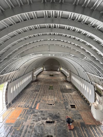 The image shows the interior of a disused airplane fuselage, likely a Boeing 747, with its interior stripped bare. The ribbed, grey metal structure of the ceiling and walls is visible, with rows of oval windows lining the sides. The floor is made of dark, worn wood panels with visible markings and some orange discoloration, suggesting previous use or damage. There are several access hatches in the floor. At the far end of the fuselage, the circular shape of a bulkhea is visible. In the foreground, on the floor, lies a black helmet on a red and beige object. The scene appears to be located in an open, possibly outdoor, environment given the natural light and greenery visible through the open doors on the left side. The location is identified as being in Pak Nam Pran, Thailand. There are no people visible in the image.