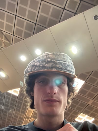 A selfie from a low angle captures a person wearing a patterned cap with a brown brim. The person has curly hair and is dressed in a dark top. Overhead, there is a grid-patterned ceiling with evenly spaced lights. The image is slightly rotated, giving a tilted perspective of the surroundings.