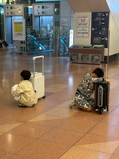 The image shows two young boys sitting on the floor of a modern airport terminal in Ota, Japan. The floor is tiled with a patterned beige and orange material.
The boy on the left is wearing a light brown jacket and is crouched down with a white bundle in his lap. Next to him is a closed, off-white suitcase with a retractable handle extended.
The boy on the right is leaning his head on a closed, black carry-on suitcase. He is wearing a black and white patterned tracksuit and appears to be sleeping. The suitcase has wheels and a handle.
In the background, there are signs with Japanese and English text, indicating "Departures" and "Taxi." There are also escalators and glass barriers, typical of an airport environment. A recycling bin unit is visible in the mid-ground. The overall lighting suggests an indoor, well-lit space.