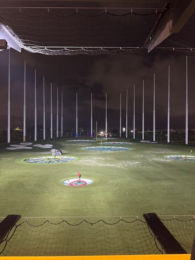 The image displays the Topgolf driving range in Miami Gardens, Florida, United States, at night. The scene is illuminated by artificial lighting under an overcast, dark sky. From an elevated vantage point within a golf bay, black protective netting is visible across the immediate foreground. The midground features a large, green artificial turf field scattered with numerous white golf balls. Embedded within the turf are multiple circular target areas of various sizes, some containing nets, while others are marked with flags (one red, one yellow). A prominent yellow American football goalpost stands centrally towards the back of the range. To the left-midground, a dark, irregularly shaped object with yellow markings rests on the turf. The background is characterized by a linear arrangement of approximately 15 to 20 tall, white, illuminated poles. Several of these poles, particularly on the left side, display blue and purple/pink lighting. Distant foliage and faint structural outlines are discernible behind the poles. No individuals are present in the scene.