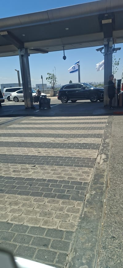 The image shows a street scene with a covered walkway. In the foreground, there is a brick-patterned pavement. A black car is parked under the walkway, and an Israeli flag is visible. Further down the walkway, a white car is parked. Several people are seen standing, including one person next to the black car and another near the white car. Luggage is also visible near the black car. The scene appears to be outdoors, with a clear blue sky indicating daytime. The sun casts shadows on the pavement, suggesting it is either morning or afternoon. The presence of the Israeli flag and the general architectural style might suggest a location in Israel, though this is not explicitly stated. The overall mood seems neutral, depicting a common street or parking area. No legible text is clearly visible in the image.