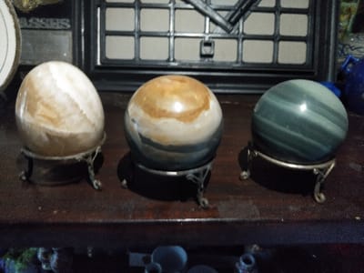 The scene, located indoors in Steenbergen, Netherlands, displays three polished mineral spheres arranged horizontally on a dark, polished wooden surface. From left to right: The first sphere, appearing ovoid, is light brown and white with swirling patterns, supported by an ornate, tarnished silver-colored metal tripod stand. The central sphere is spherical, displaying concentric bands of tan, white, and dark grey-blue, resting on a black circular base which is itself supported by a simpler dark metal tripod stand. The third sphere is spherical, featuring horizontal bands of dark teal and lighter green, and sits on an ornate, tarnished silver-colored metal tripod stand matching the first. The background is out of focus, revealing a dark rectangular grid structure behind the spheres. To the far left, a partial round white object with a gold rim displays the partial text 'ASSES'. To the far right, a partial blue ceramic object is visible. No individuals are present, and the scene is static.