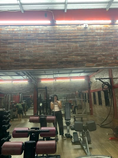 The image shows the interior of a gym, featuring a large mirror reflecting the space. In the foreground, there are several weightlifting machines. To the right of the image, a black rope is suspended from the ceiling. The image is taken from the perspective of a seated position, looking straight ahead. There is a person in the reflection of the mirror standing in the background. The person is wearing a pink shirt and black pants and appears to be looking at their phone. The room appears to be dimly lit. The gym is located at Latitude: -7.568 and Longitude: 110.757 in Kecamatan Kartasura, Indonesia.