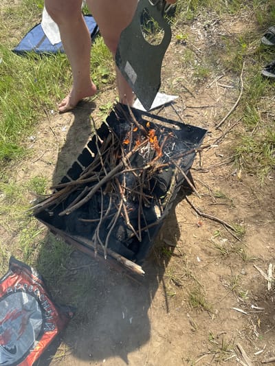 The image depicts an outdoor scene, likely in a natural setting, with a focus on a barbecue grill. A metal barbecue grill, filled with charcoal and burning wood, is positioned on the ground. Several dry sticks and branches are arranged within the grill, with flames and smoke emanating from them. To the left of the grill, a person's bare legs and feet are visible, suggesting they are standing. The ground around the grill is a mix of dirt and dry grass, with scattered twigs and debris. In the lower left corner, a red and silver bag, possibly containing charcoal or other barbecue supplies, is partially visible. The overall impression is of a casual outdoor gathering, likely for a barbecue. The location is identified as Pravda, Russia.