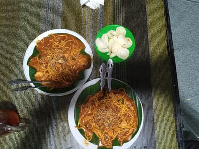 The image captures an overhead view of a dining scene in Keudealuerheung, Indonesia. Two servings of noodles are prominently displayed on a textured, striped table surface. Each serving is presented in a white, round ceramic bowl or plate, lined with a green banana leaf. The noodles are yellow and coated in a thick, reddish-brown sauce, topped with a generous sprinkle of fried shallots. Visible ingredients within the sauce include small pieces of meat, possibly chicken, and bean sprouts. Each plate is accompanied by two ornate silver-colored spoons. To the upper right, a small, vibrant green plastic bowl contains a pile of white, irregular-shaped krupuk (crackers). In the lower-left corner, a clear glass mug holds a reddish-brown beverage, likely iced tea, with a visible slice of citrus fruit. The table surface is a tightly woven rug or mat featuring vertical stripes in shades of olive green, beige, and dark brown. A crumpled white napkin is situated near the top center of the frame. The lighting suggests an indoor setting, and no individuals are present in the frame.
