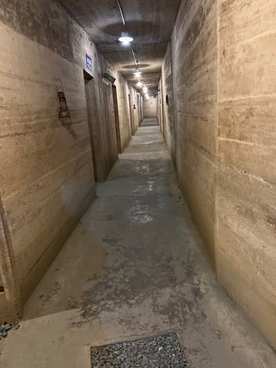 This media file presents a low-angle, deep perspective view looking down or along a long, narrow concrete passage or shaft in Trân Châu, Vietnam. The scene is dominated by raw, utilitarian concrete surfaces forming the walls, floor, and ceiling, which show signs of wear, staining, and textural variations.
On the left side of the passage, a series of bright, circular lights with white covers are mounted on the concrete wall, providing illumination. Exposed electrical conduit runs horizontally along the wall, connecting these lights. The floor of the passage appears to be constructed with multiple concrete levels or steps that recede into the distance, creating a sense of depth and a descending path.
Towards the foreground on the left, a blue rectangular warning sign with white text is affixed to one of the concrete ledges. The word "WARNING" is clearly visible at the top of the sign, followed by additional lines of safety-related text that are illegible due to distance. On the right side of the passage, near the top, a section of the concrete wall is filled with small, grey angular gravel or crushed stone. No people are visible in the image. The artificial lighting suggests an enclosed, possibly subterranean, environment, indicative of infrastructure, a utility tunnel, or a deep accessway.
