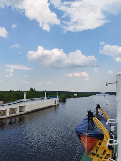 The image depicts a canal scene, likely taken from a boat or ship. The sky is a clear blue with scattered white clouds. On the left is a long, white concrete structure with architectural details and what appear to be decorative posts on top. Vegetation, possibly trees and shrubs, lines the canal's edge beyond the structure. The canal itself is wide, reflecting the sky and surroundings. In the distance, other boats or ships are visible. On the right, a portion of the boat or ship from which the photo was taken is visible, including a lifeboat covered with a blue tarp and supported by yellow cranes. The inscription on the lifeboat reads "Евгений Орловский, Котлас," indicating the vessel's name and home port. Although Karmanovo, Russia, is mentioned as the location, it is not visibly discernible from the image alone without additional information.