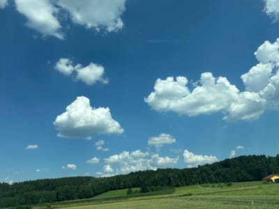 This media file depicts a reflection of a landscape onto a body of water. The landscape features a green field and a line of trees. A small yellow building is visible in the upper left corner of the landscape. The sky is blue with fluffy white clouds. The overall scene suggests a clear day, likely in the afternoon given the bright sunlight casting reflections. The location is identified as Halfing, Germany. No people, text, or specific activities are discernable. The image is oriented such that the landscape appears at the top, and its reflection in the water is below, creating a symmetrical effect.