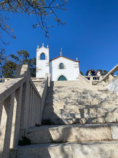 The image shows the Sanctuary of Monte da Saúde in Esposende, Portugal. A white chapel stands at the top of a long stone staircase. A group of people is visible at the top of the stairs near the chapel. The sky is clear and blue. Trees are visible in the foreground and behind the chapel. A stone railing lines the staircase.