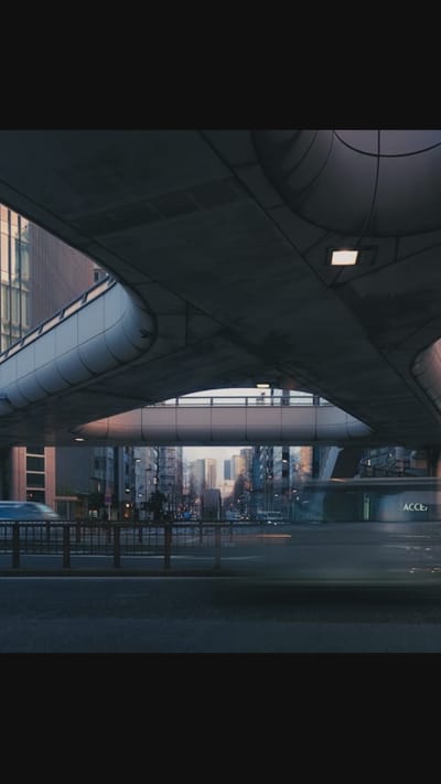 The image shows a low-angle view of an elevated pedestrian walkway or overpass in a city, likely during dusk or dawn. The walkway is constructed of concrete and metal, with a curved, modern design. Below the walkway, a street is visible with traffic, indicated by blurred streaks of light from vehicles. Buildings line the street, appearing to be a mix of commercial and residential structures. The sky transitions from a muted pink and orange near the horizon to a deep blue overhead. A sign with the partial text "ACCE" is visible on one of the buildings. The overall atmosphere is one of urban activity and infrastructure. The location is not Oreshki, Russia.