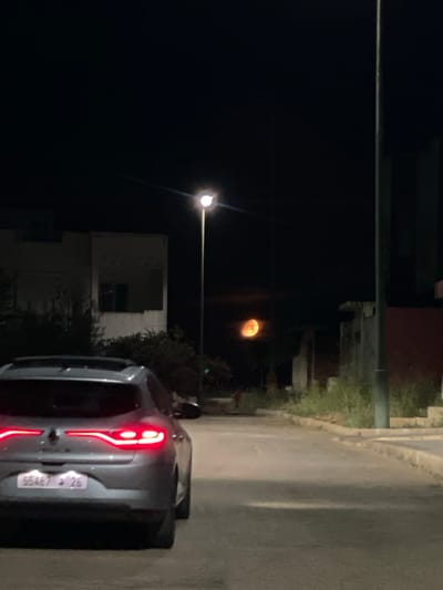 A vertical, dimly lit nighttime shot shows a car parked on a street in Dar Oum Soltane, Morocco. The rear of the car, facing the camera, shows illuminated taillights and a license plate. A street lamp is visible behind the car, with buildings and vegetation in the background. Above the buildings a full, orangish moon is visible. The street is to the right, and the overall scene is dark with limited illumination.