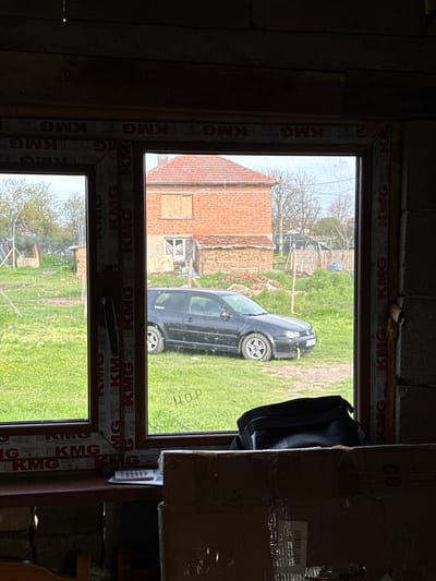 The image presents a view looking out from inside a dimly lit room, possibly under construction or renovation, through a large PVC window. The interior walls appear to be constructed from concrete blocks and unfinished wooden planks. The window frame is brown, and visible branding indicates it is a "KMG Austrian technology system."
Through the window, an outdoor scene unfolds in daylight. A grassy, somewhat unkempt field or yard occupies the middle ground. A dark-colored car, possibly a compact SUV or hatchback, is parked on the grass, facing towards the left side of the frame (when correctly oriented). In the background, a brick building with a red tiled roof is visible, suggesting a house or outbuilding, with one of its windows appearing to be boarded up. Sparsely leaved trees are scattered across the landscape.
Inside the room, several objects are visible near the window frame. A black bag or case hangs from the upper right side of the window. To the right, a large cardboard box partially obstructs the view. A small white remote control is attached to the window frame on the right. Text on the cardboard box includes "16803025BK" and "QTY 30.40LBS". The outdoor scene, featuring the brick building and green landscape, is consistent with a rural or semi-rural setting in Straldzha, Bulgaria. No specific activity or people are depicted, giving the scene a quiet, static quality. The light suggests it is daytime, with no strong shadows indicating harsh sunlight.