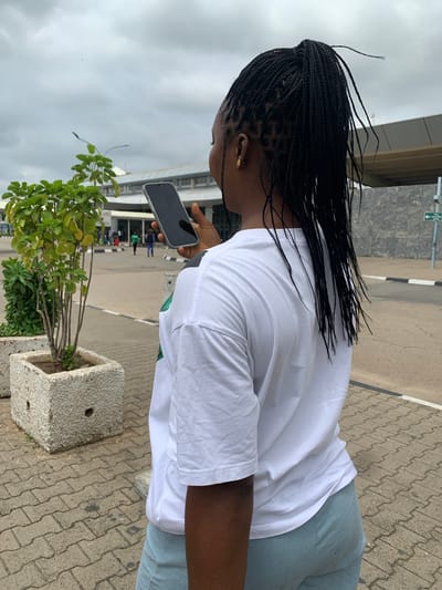 The image shows a person from a back and side profile, standing outdoors on a paved surface. The setting appears to be an airport or a public building entrance in Gwaska, Nigeria, characterized by a large structure in the background and a paved forecourt. The sky is overcast.
The main subject is an individual with dark skin, wearing a plain white short-sleeved t-shirt and light blue or grey pants. Their long, dark hair is styled in numerous thin braids, some of which are slightly dishevelled, suggesting a light breeze. A small, golden earring is visible on their right ear. The person is holding a dark-screened smartphone with a light-colored frame in their right hand, appearing to be focused on the device.
The ground is covered with grey interlocking paver stones. To the right of the person, a grey concrete planter containing small green leafy plants is visible. In the background, a large, modern-looking building with a grey facade and windows can be seen. A road or pathway with white painted lines runs adjacent to the paved area. A green directional sign with a white arrow pointing to the right is partially visible in the upper background. The time of day is daytime, under a cloudy sky. No other specific activities or identifiable text are visible.