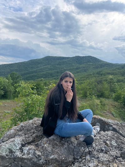A young woman with long, dark hair sits on a rocky outcrop overlooking a densely forested hillside. She is wearing light blue jeans, grey and white sneakers, and a black jacket. She holds a lit cigarette in her left hand, close to her mouth. The background features a cloudy sky, a steep, tree-covered mountain, and a few smaller trees growing near the edge of the rock where she sits. The location is likely in Kamchia, Bulgaria, based on the presence of the characteristic landscape. The scene appears to be an outdoor setting on a somewhat overcast day.