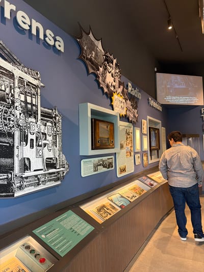 The image shows a museum exhibit in Tampico, Mexico, focused on the history of media. "Prensa" (Press), "Radio," and "Televisión" are labeled on the wall, accompanied by related images and objects. There's a newspaper printing press illustration, radio displays, and television sets. Text panels provide information about these media forms in Tampico. A man wearing jeans and a plaid shirt is examining the television display. The exhibit includes newspapers from Tampico from 1910 to 1950, and a description of events carried out in the city. There is also a botonera of an old print.